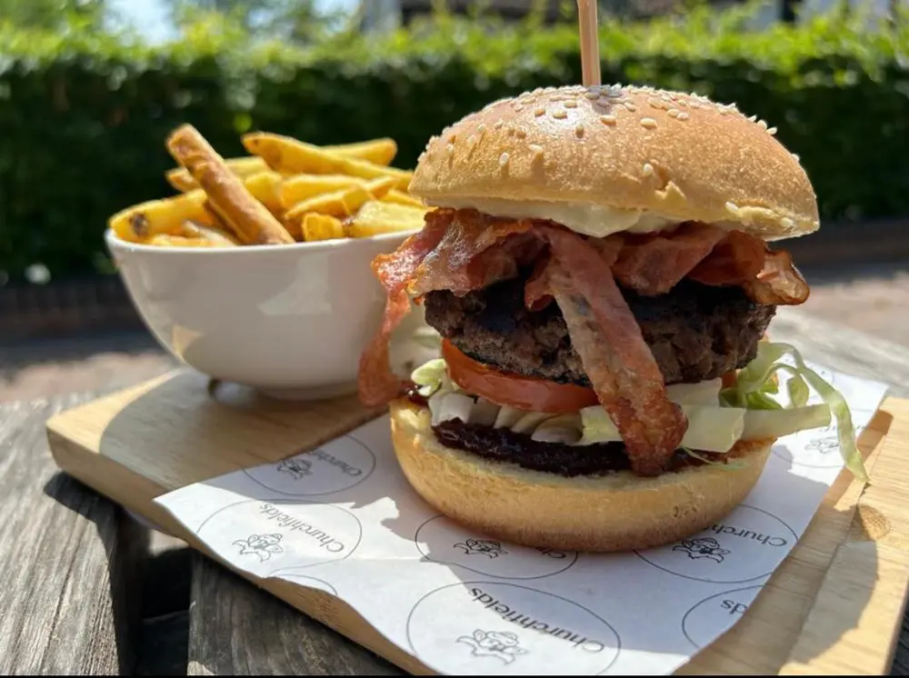 Close up of Large burger and bowl of chips served outside with hedge background