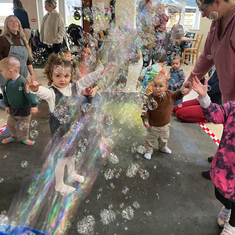 Small children and toddlers playing with bubbles coming out of bubble machine with mums in background.