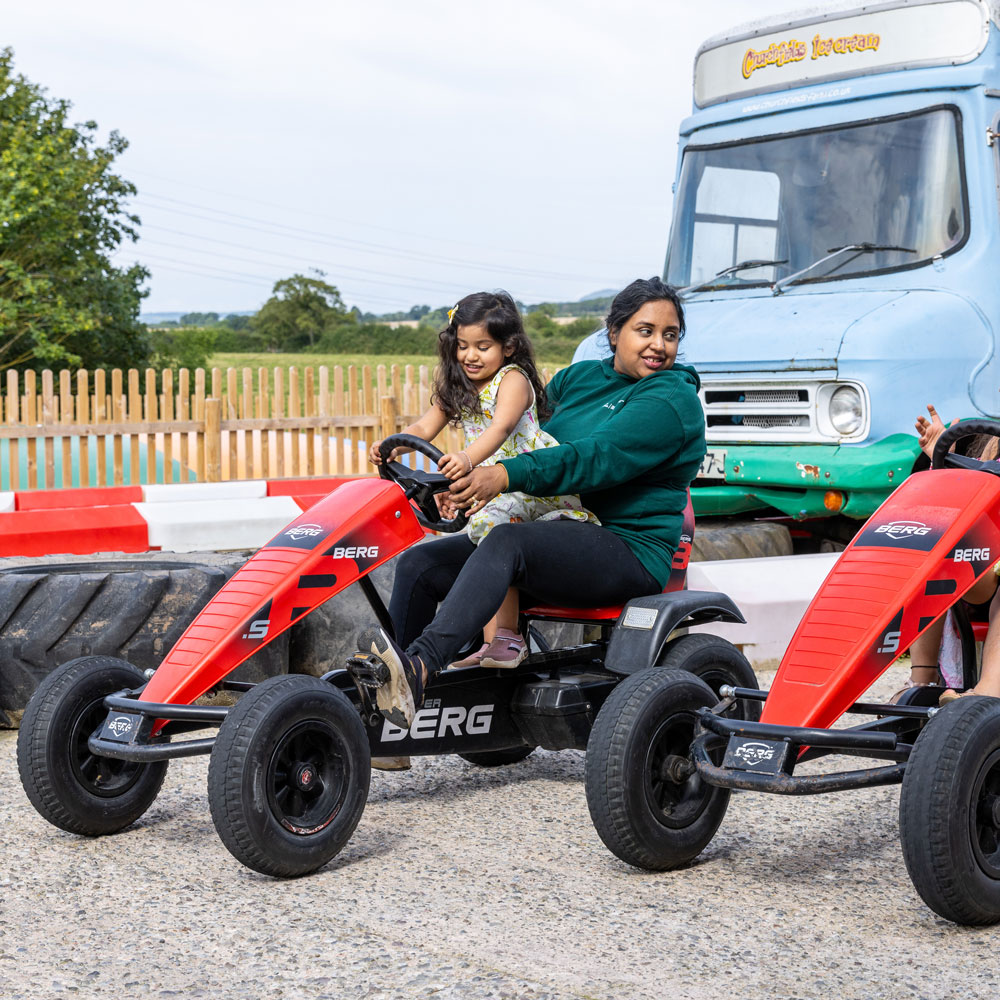 Mum and daughter on go kart as mum pedals and daughter steers on go kart track with old Churchfields Ice Cream van in background.