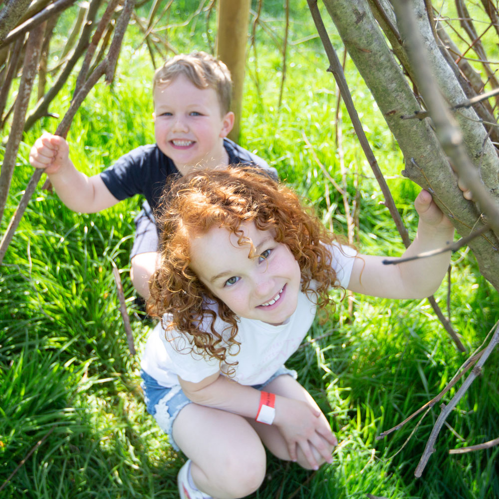 Small brother and sister smile for camera inside a den made of sticks leaning together on green grass
